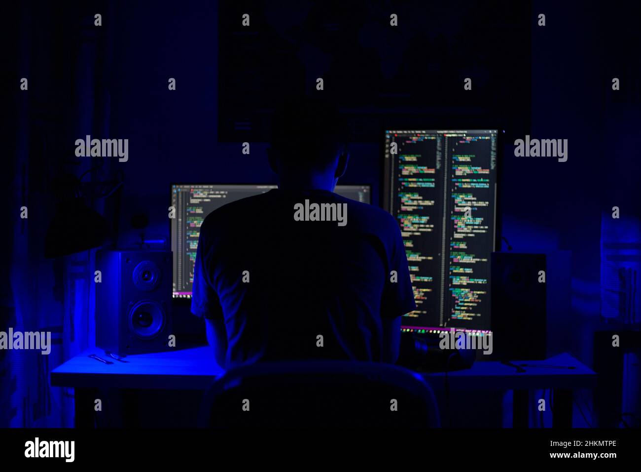 Ein Mann sitzt nachts in einem Raum an einem Tisch mit blauer Beleuchtung und Programmen an einem Computer Stockfoto