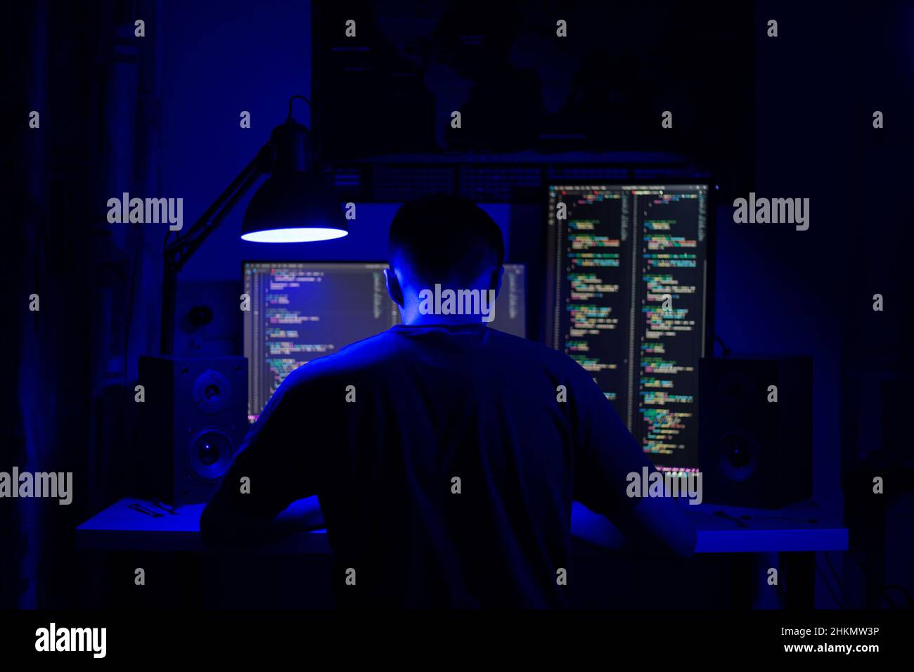 Ein Mann sitzt nachts in einem Raum an einem Tisch mit blauer Beleuchtung und Programmen an einem Computer Stockfoto