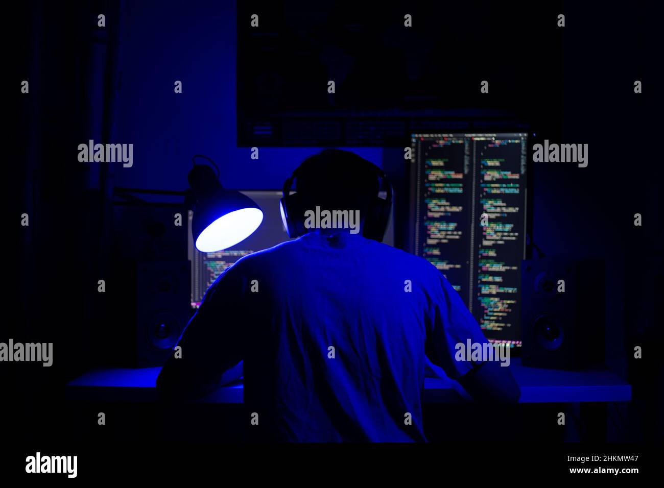 Ein Mann sitzt nachts in einem Raum an einem Tisch mit blauer Beleuchtung und Programmen an einem Computer Stockfoto