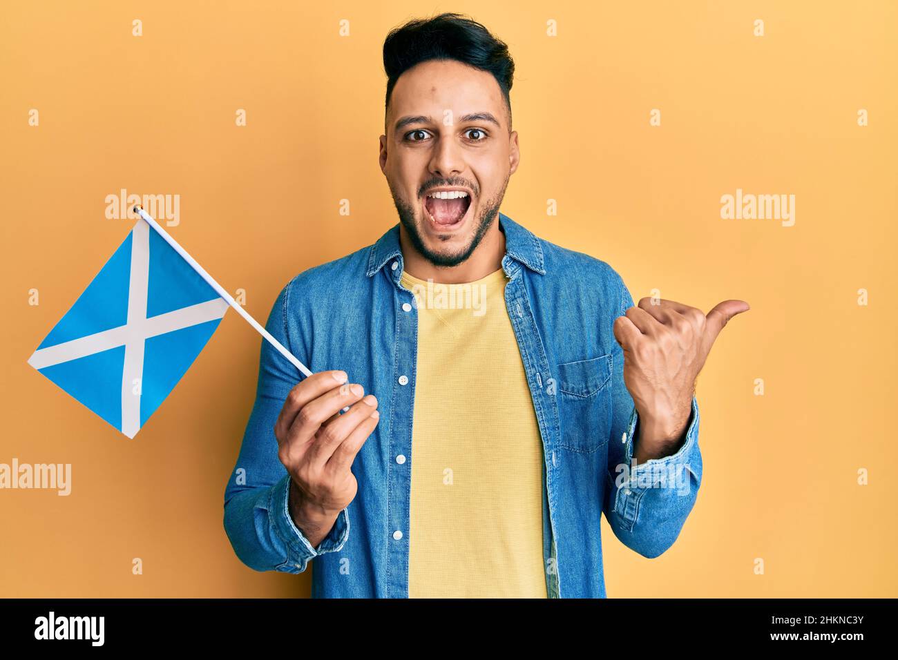 Junger araber, der die schottische Flagge hält und den Daumen nach oben zur Seite zeigt, lächelt glücklich mit offenem Mund Stockfoto