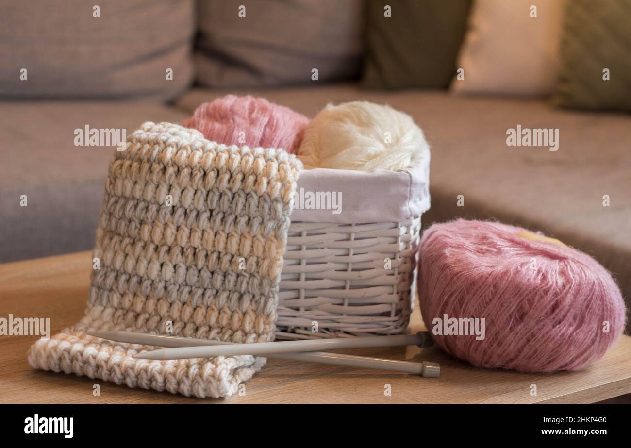 Rosa und weiße Wollkugeln in einem weißen Korb mit Stricknadeln und einem handgefertigten Strickschal Stockfoto