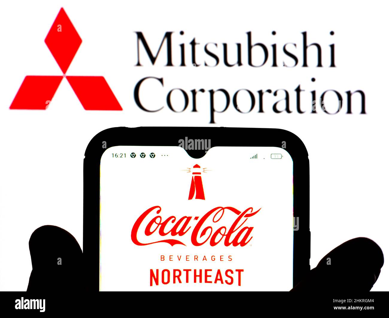 In dieser Abbildung wird das Coca-Cola Beverages Northeast-Logo auf einem Smartphone-Bildschirm mit einem Mitsubishi Corporation-Logo im Hintergrund angezeigt. Stockfoto