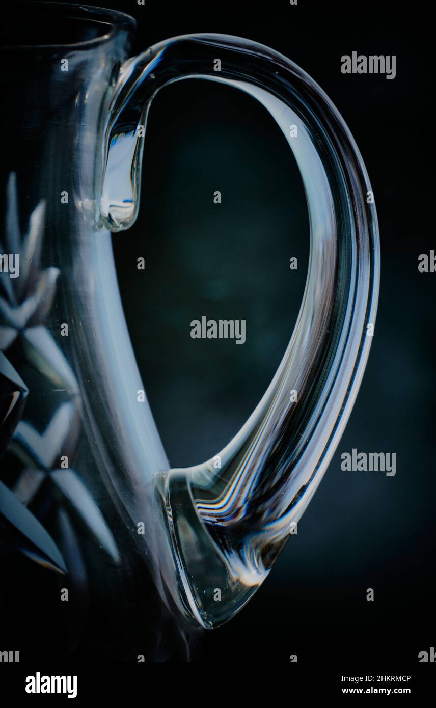 Exquisit geschwungener Griff an alter, geschnittener Glaskanne. Die Griffwölbung bricht Farben vom Hintergrund, einschließlich Blau- und Waldgrün Stockfoto