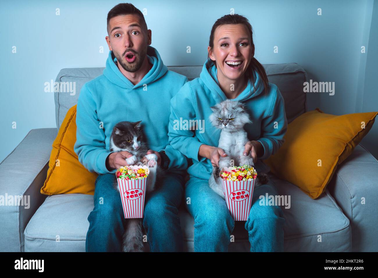 Ein junges, glückliches Paar mit Popcorn und lustigen Katzen, die auf dem Sofa sitzen und Comedy-Filme ansehen und die Zeit zu Hause genießen Stockfoto