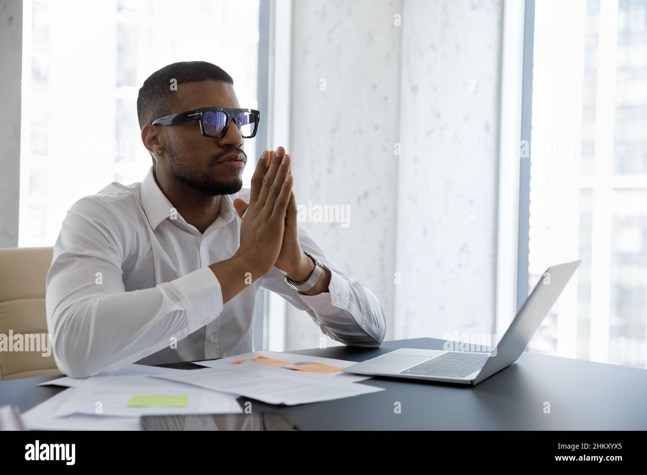 Friedlicher, ernsthafter junger afroamerikanischer Geschäftsmann, der am Arbeitsplatz meditiert und betet Stockfoto