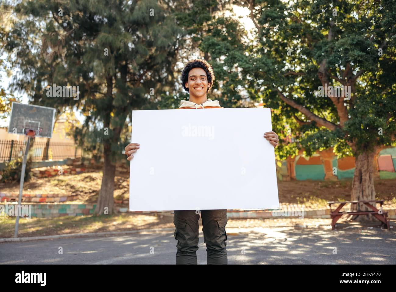 Lächelnder Teenager-Aktivist, der ein leeres Banner im Freien zeigt. Selbstbewusster Teenager, der tagsüber ein weißes Plakat hält. Teenager, der die Kamera ansieht Stockfoto