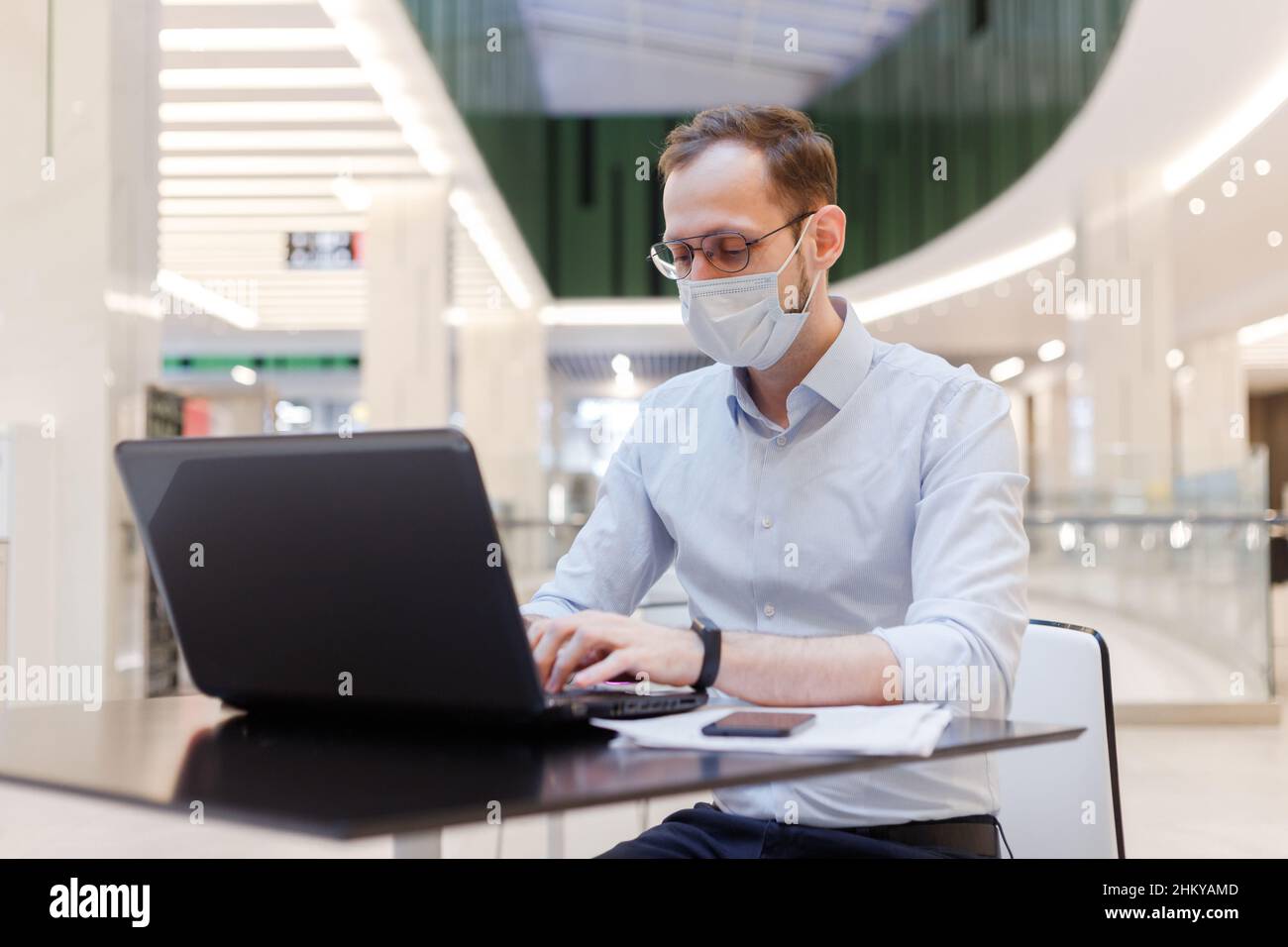 Angestellter mit Gesichtsmaske, Mann, der an einem öffentlichen Ort an einem Laptop arbeitet Stockfoto