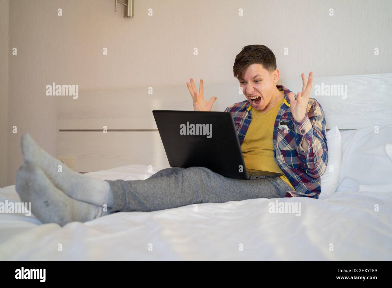 Frustrierter, ärgerlicher Mann, der zu Hause im Büro auf dem Bett sitzt und einen kaputten laptop ansieht, empfindet Wut und Empörung. Ängstlicher Mann hat Probleme Stockfoto