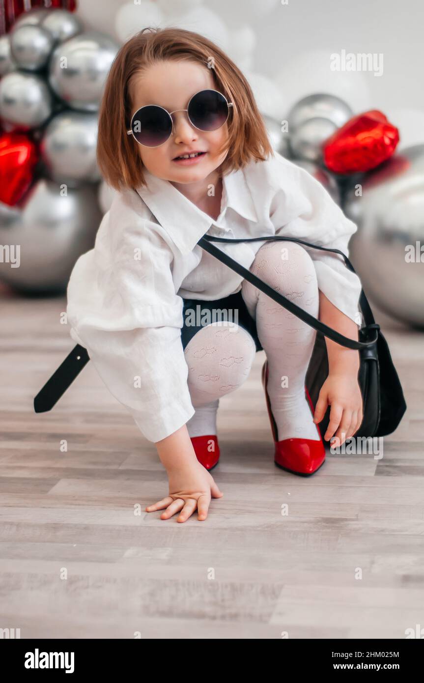 Ein modisches Baby in Sonnenbrillen versucht an den roten Schuhen ihrer Mutter. Mode und moderne Kleidung. Glitter hell metallic Spiegel Hintergrund, neue Trends V Stockfoto