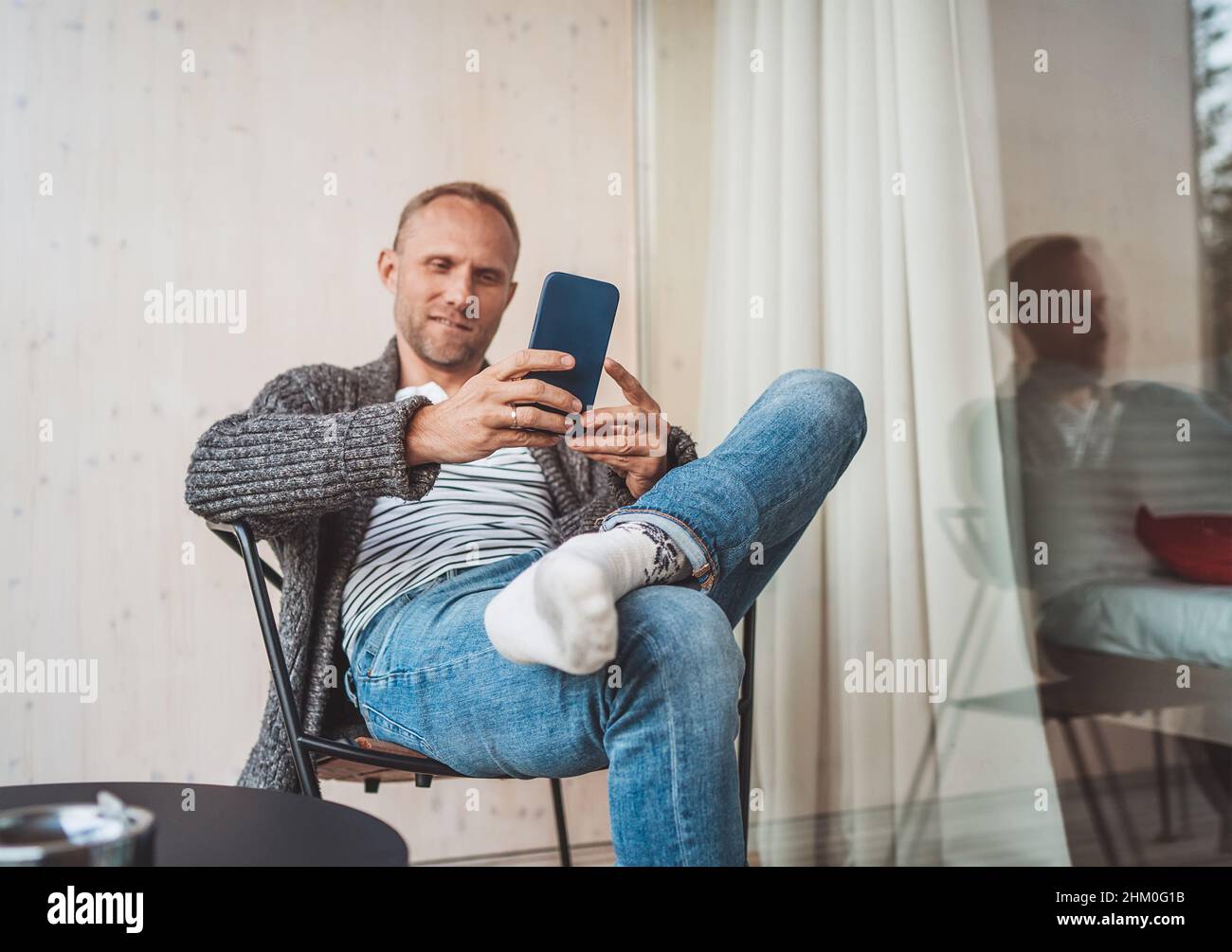 Lächelnder Mann mittleren Alters, der offene Strickjacke, Jeans und warme Socken angezogen hat und auf dem Balkon des Hauses sitzt und lächelt, wenn er ein modernes Smartphone benutzt. Tägliche li Stockfoto