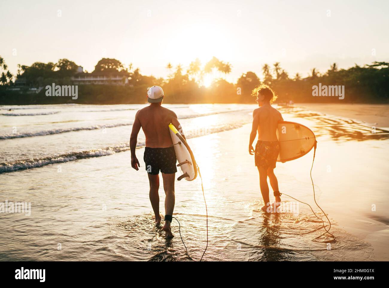 Vater mit Teenager-Sohn, der mit Surfbrettern am Sandstrand mit Palmen im Hintergrund, der von der Sonne des Sonnenuntergangs erhellt wird, entlang läuft. Sie lächeln und ha Stockfoto