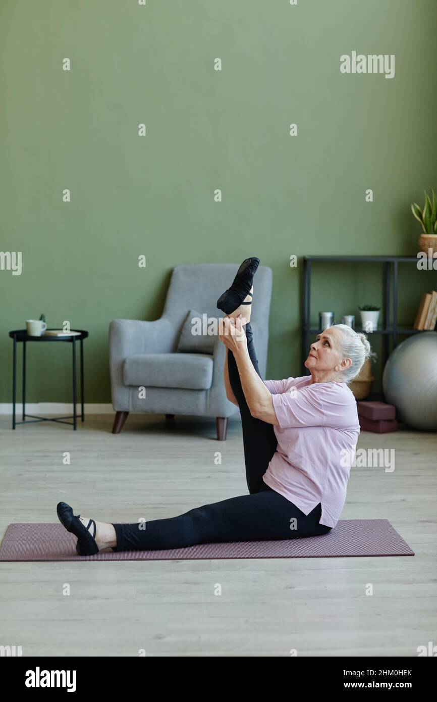Senior-Sportlerin im beigen T-Shirt und schwarzen Leggins hält und hebt das gestreckte Bein beim Training auf der Yogamatte Stockfoto