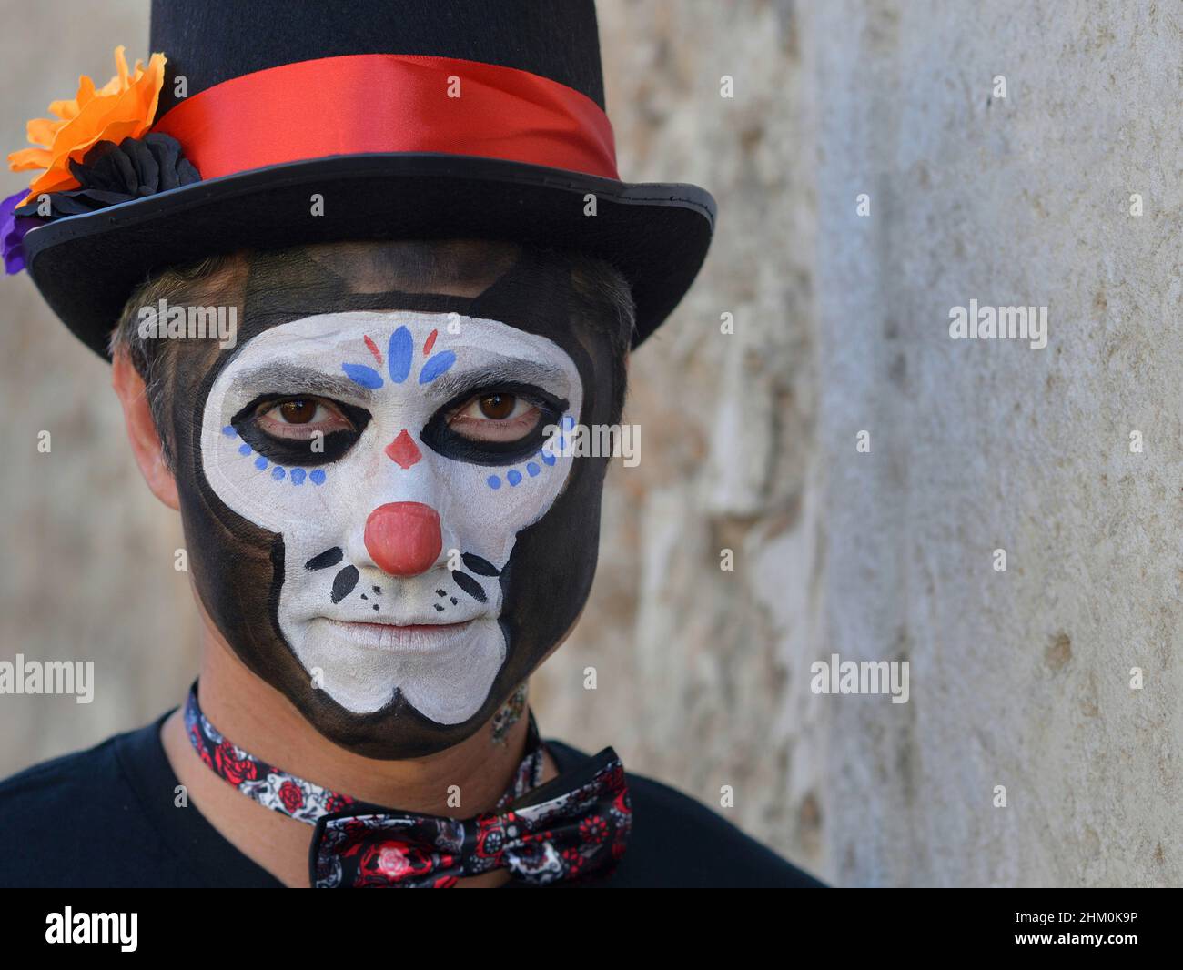 Der junge kaukasische Mann mit gruseliger traditioneller weißer Gesichtsbemalung trägt am Tag der Toten (Día de los Muertos) einen schwarzen Hut und blickt auf den Betrachter. Stockfoto