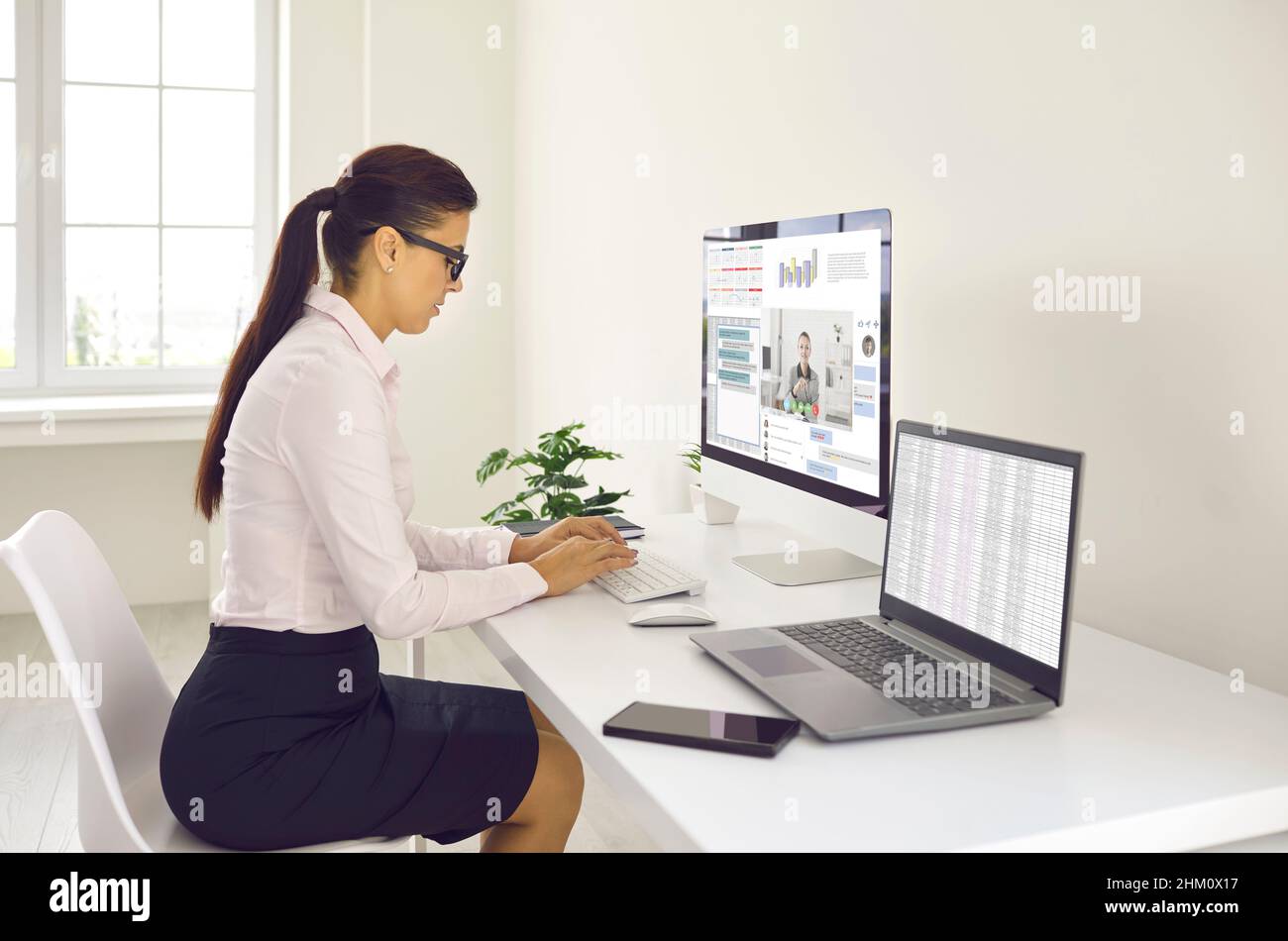 Junge Frau, die am Schreibtisch sitzt, mehrere Computer nutzt und im Arbeitschat Nachrichten mitgibt Stockfoto