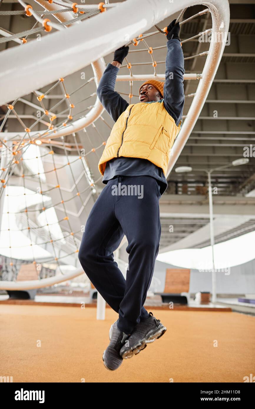 Junge afroamerikanische Athletin in Sportbekleidung, die während des Pull-up-Trainings auf dem Sportplatz am Seilnetz hängt Stockfoto