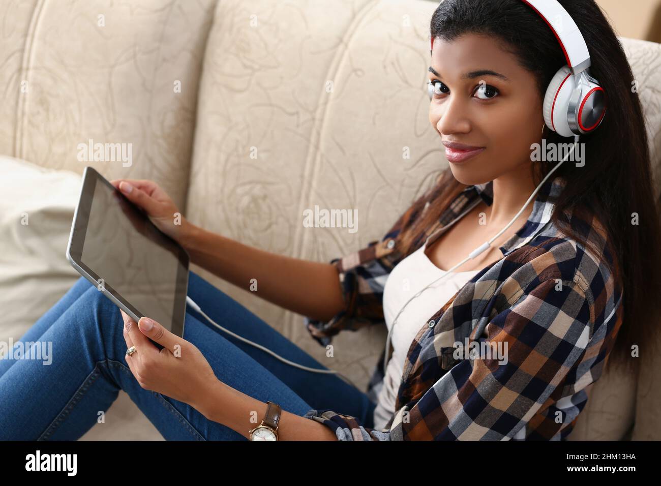 Ein dunkelhäutig geteiltes Mädchen mit Kopfhörern und Tablet sitzt auf dem Sofa Stockfoto
