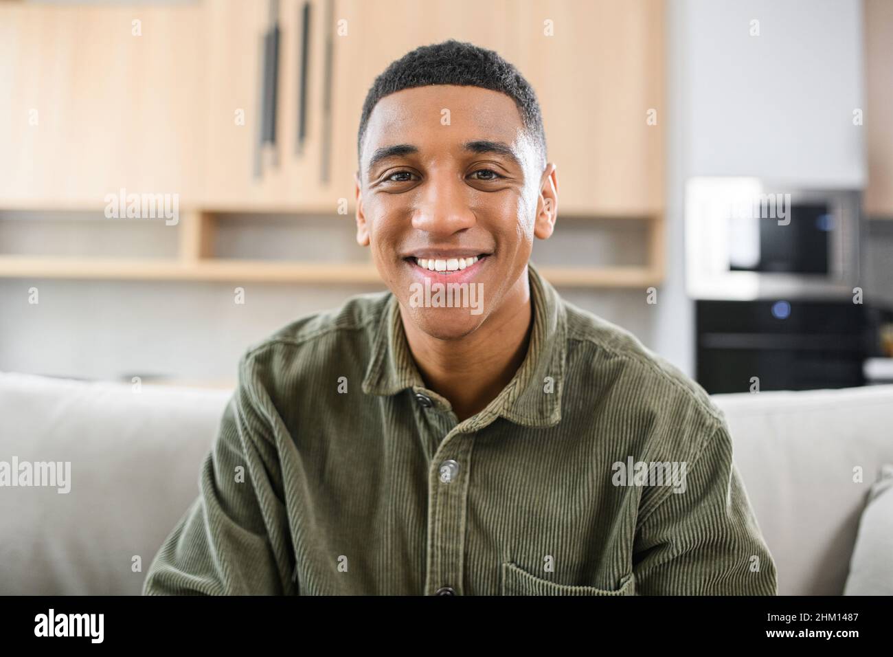 Porträt eines sorglosen jungen Mannes, der auf dem Sofa sitzt und die Kamera anschaut. Heiterer Mann in legerem Hemd in entspannter Atmosphäre, der Freizeit zu Hause verbringt Stockfoto
