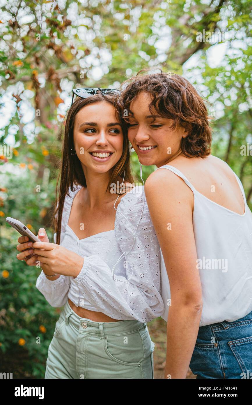 Zwei junge Teenager-Mädchen, die Musik auf weißen Kopfhörern hören und Spaß in einem Park haben. Sie lächeln einander an. Stockfoto