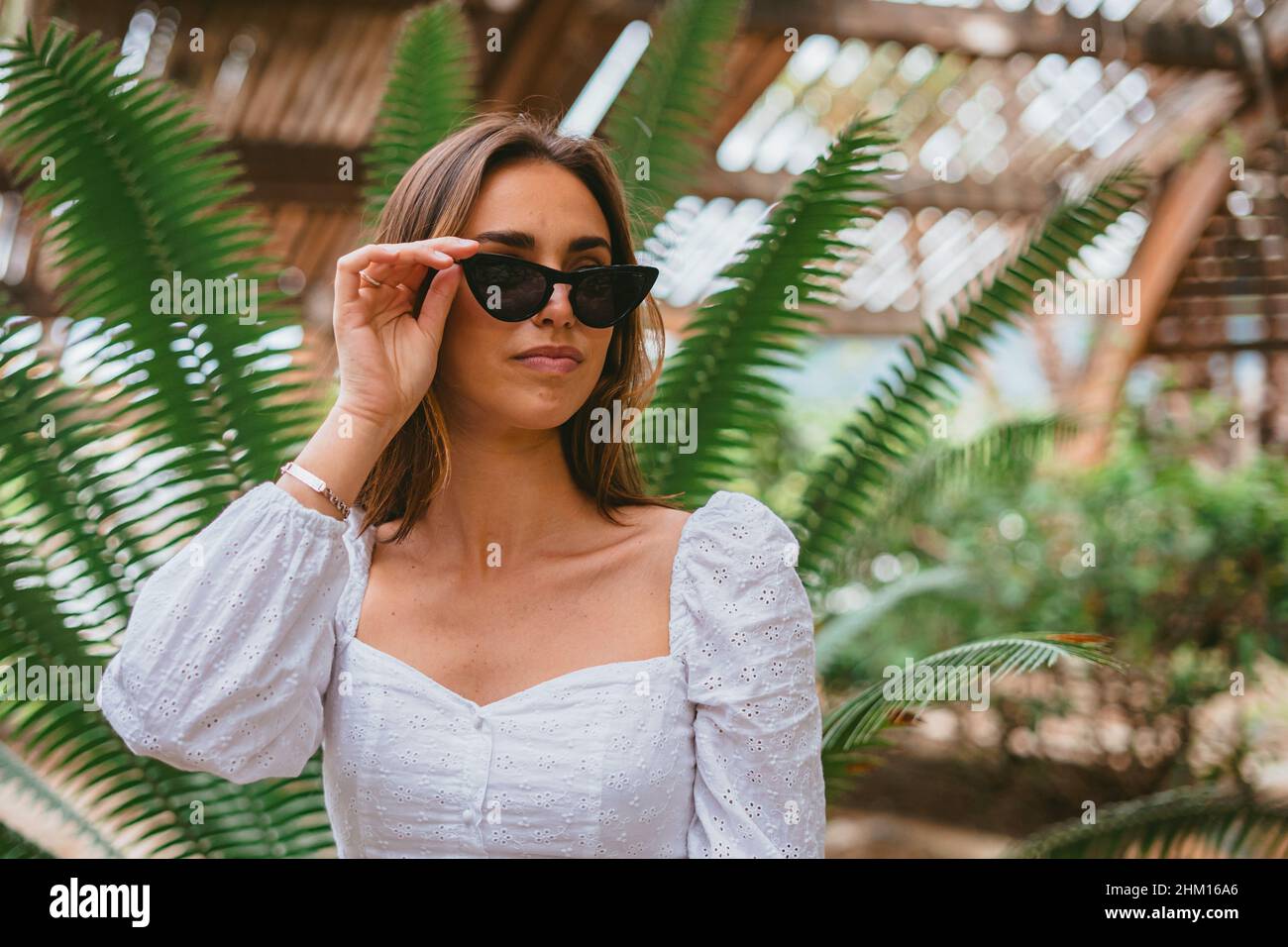 Porträt eines Teenagers, das ihre Sonnenbrille aufsetzt. Hinter ihr ist eine grüne Palme. (Platz kopieren) (Horizontale Aufnahme) Stockfoto