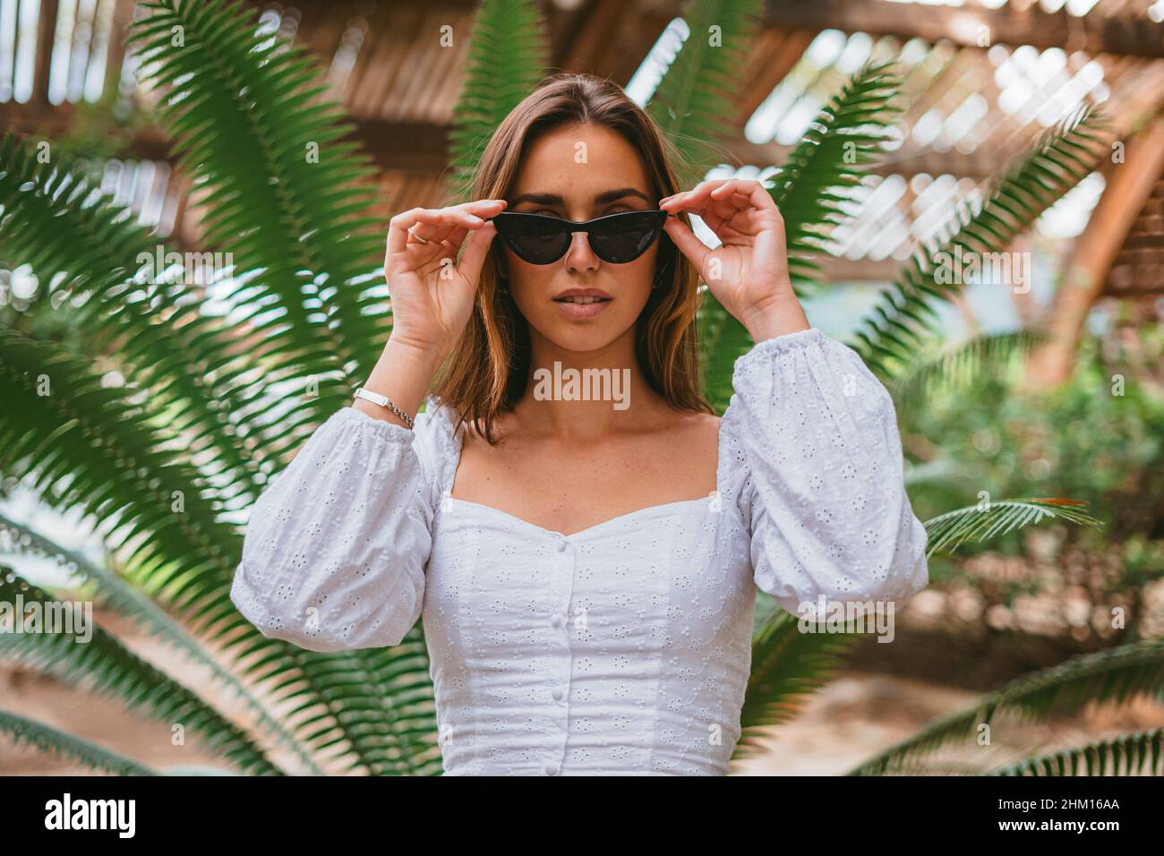 Porträt eines Teenagers, das ihre Sonnenbrille berührt. Sie ist im Park und es gibt eine Palme. (Horizontale Aufnahme) Stockfoto