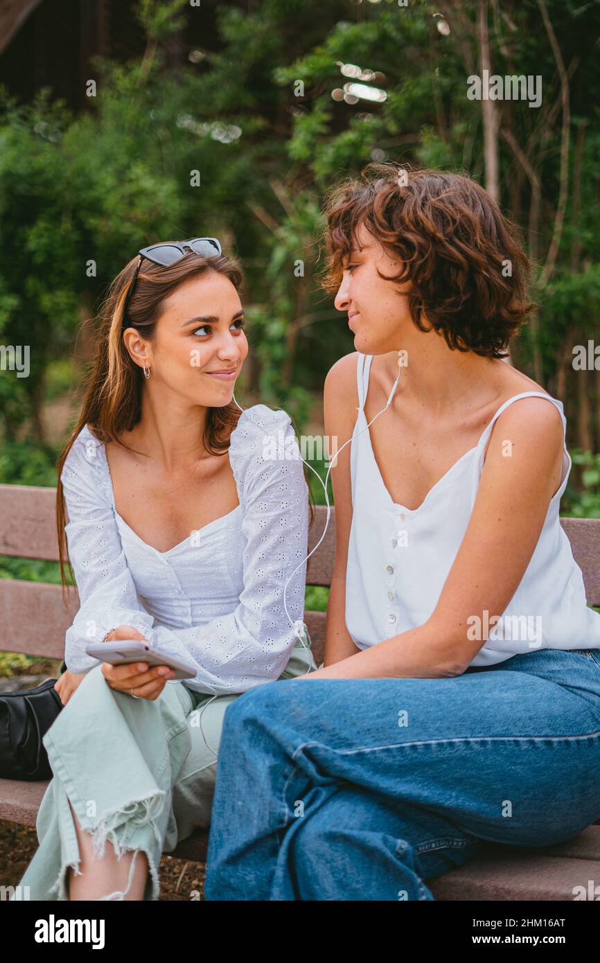 Zwei lächelnde Teenager-Mädchen, die Musik hören, während sie auf einer Holzbank im Park sitzen. Sie schauen einander an. Stockfoto