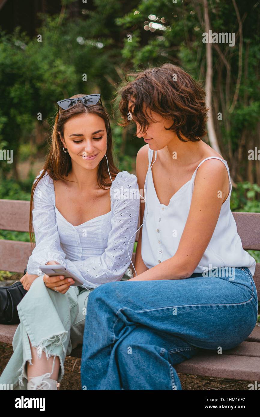 Zwei Teenager-Freunde, die am Telefon sitzen und Musik hören, während sie auf einer Bank im Park sitzen. Stockfoto