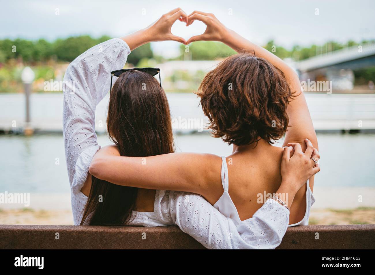 Zwei Teenager-Freundinnen, die mit ihren Händen eine Herzform machen, während sie auf einer Bank am Fluss sitzen. Stockfoto
