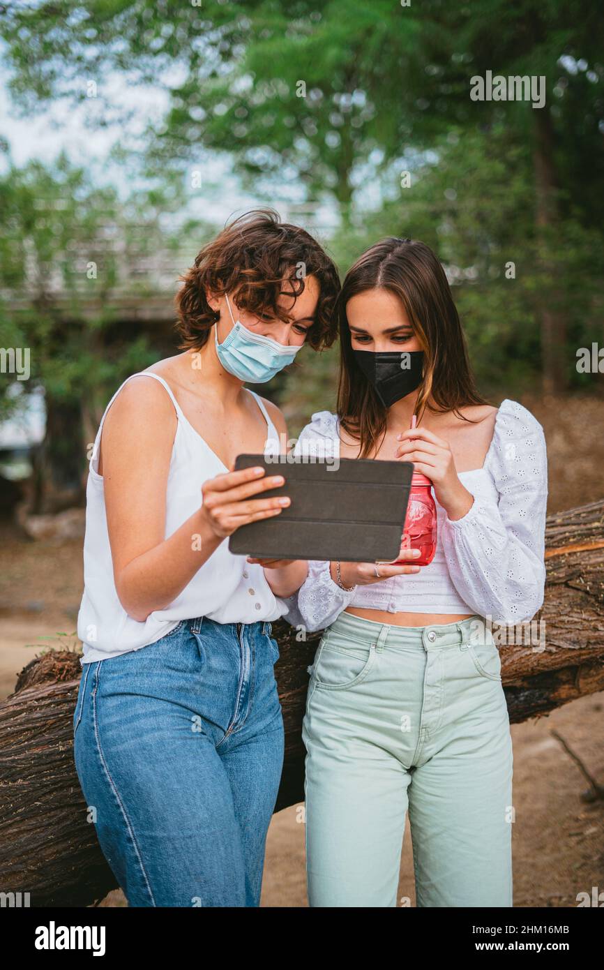 Zwei junge Frauen betrachten einen Tablet-Computer, tragen medizinische Masken und lehnen sich an einen Baum. Stockfoto