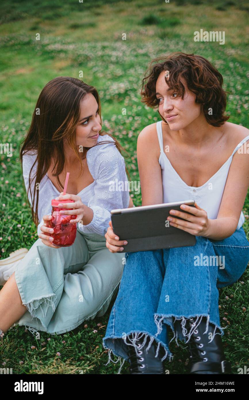 Zwei Freunde, die sich gegenseitig anguckten, während sie auf einem Tablet-Computer in sozialen Medien nachschauten. Sie sitzen auf dem Gras. Stockfoto