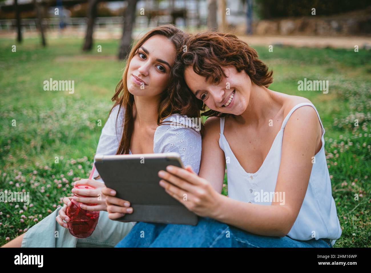 Zwei Freundinnen schauen sich im Park ein Video an, während eine von ihnen einen Smoothie trinkt Stockfoto