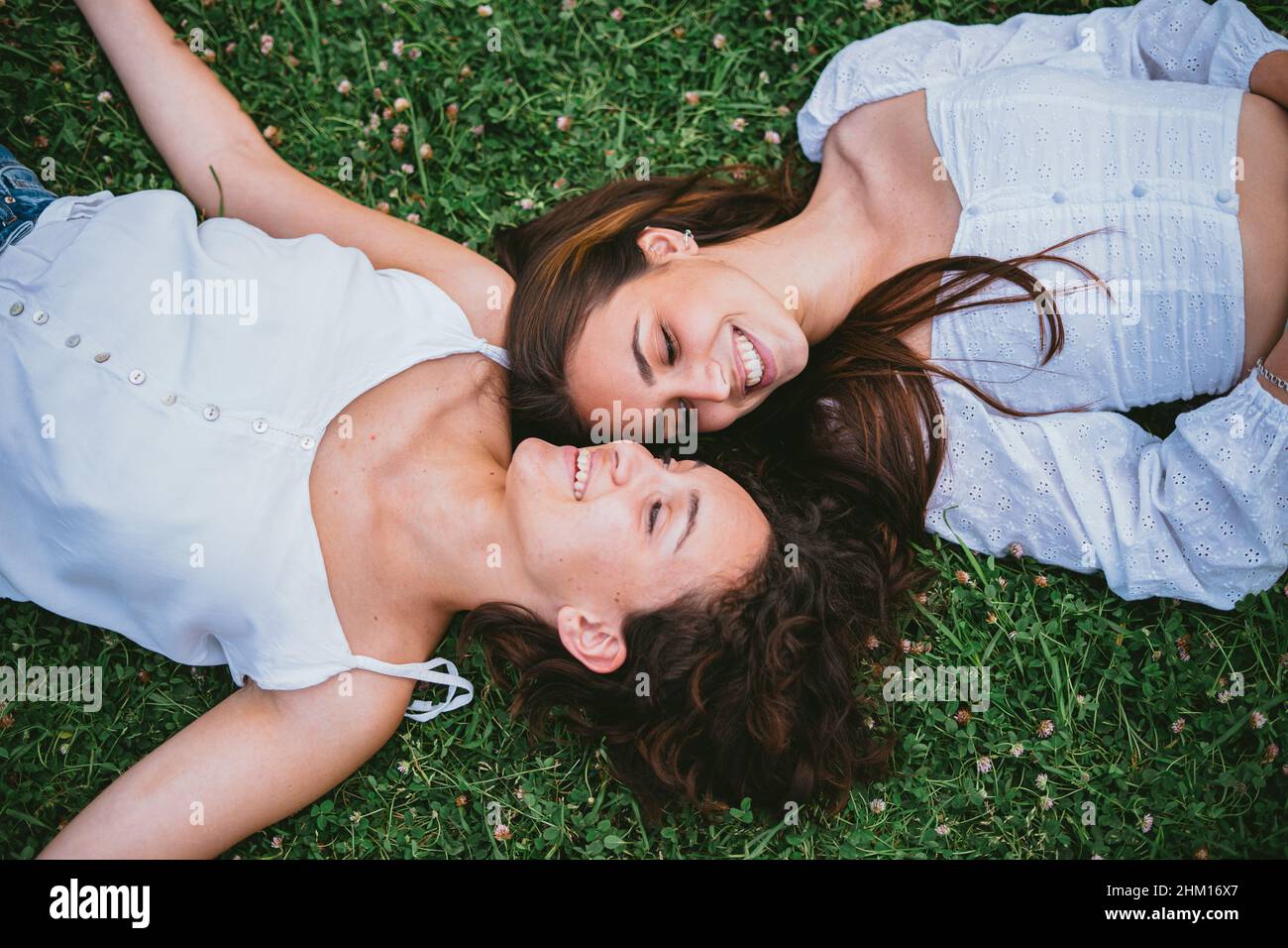 Zwei Teenager-Mädchen lächeln und blicken einander an, während sie sich in einem Park auf Gras niederlegen. Sie wirken entspannt. Stockfoto