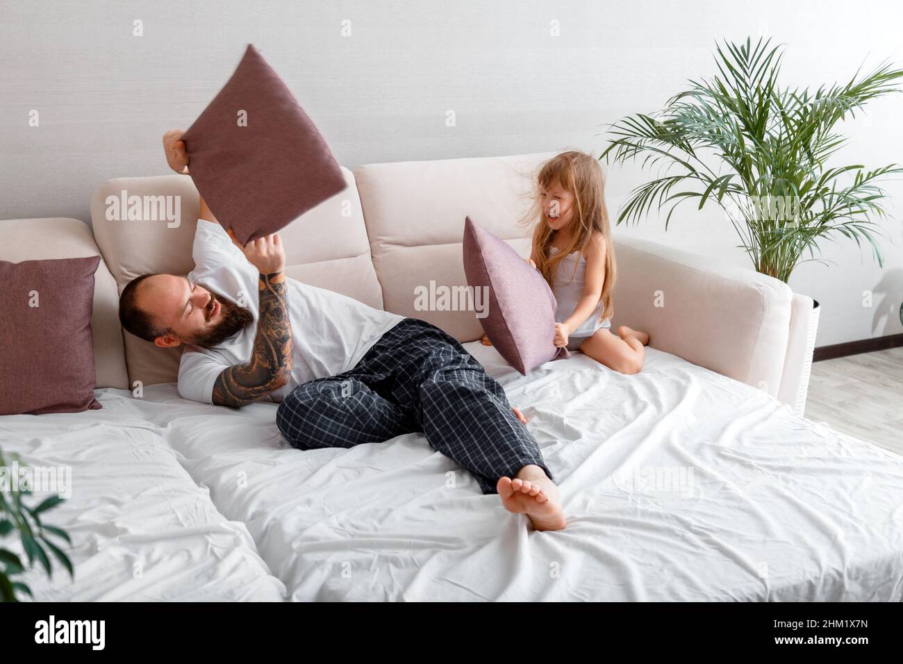 Vater und Tochter spielen im Schlafanzug im Bett gemeinsam im Kissenkampf. Kahlbärtiger Kaukasischer Mann in Tattoos und zarte kleine Tochter Stockfoto