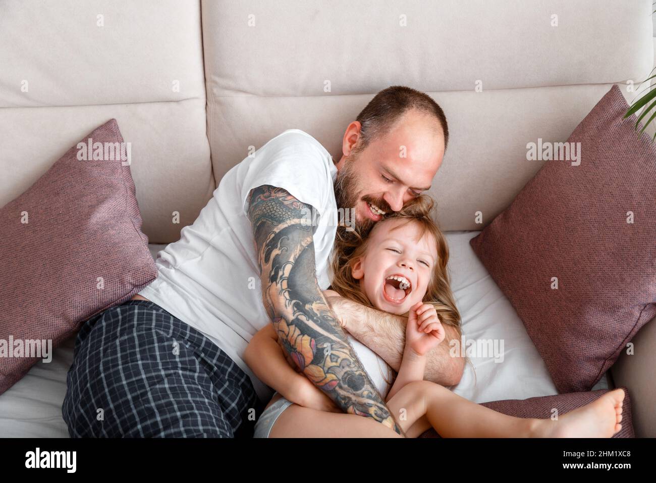 Vater spielen mit Kind Kind Mädchen verbringen Zeit zusammen. Kahlbärtiger Kaukasischer Mann mit Tattoos kitzelt seine zarte kleine Tochter. Mädchen lacht spielen Stockfoto