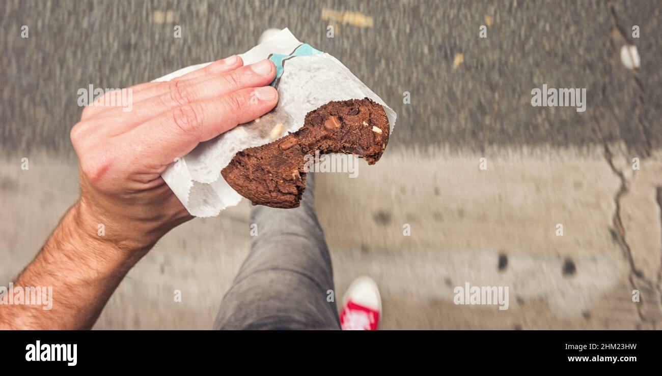 Mann, der auf dem Bürgersteig in der Stadtstraße mit gebissenen Schokoladenkekchen in der Hand geht, aus der Perspektive. Stockfoto
