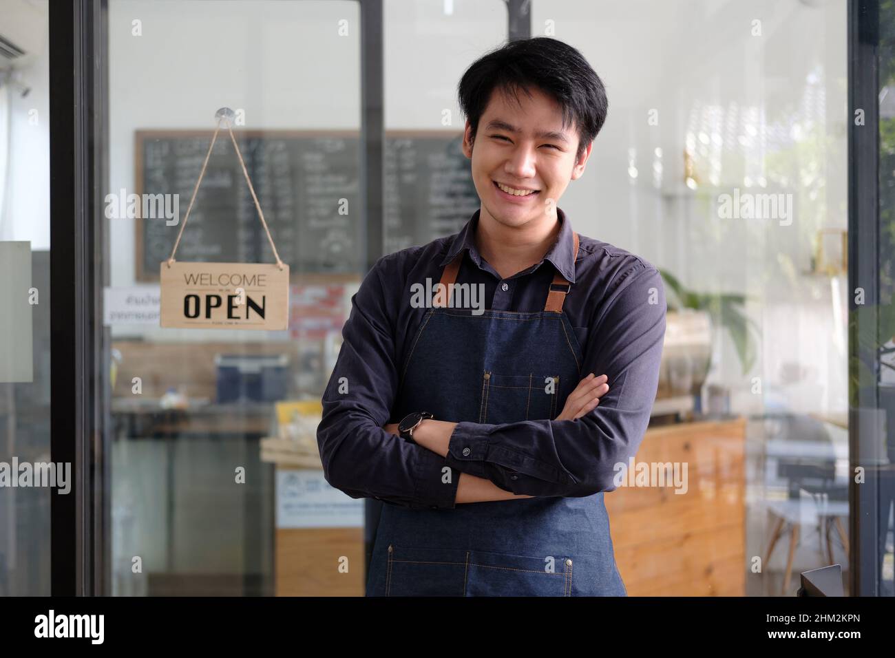 Fröhlicher junger, asiatischer Barista, der auf der Schürze ein Tablet hält und vor der Tür des Cafés mit offenem Schild steht. Unternehmer Startup SME Stockfoto