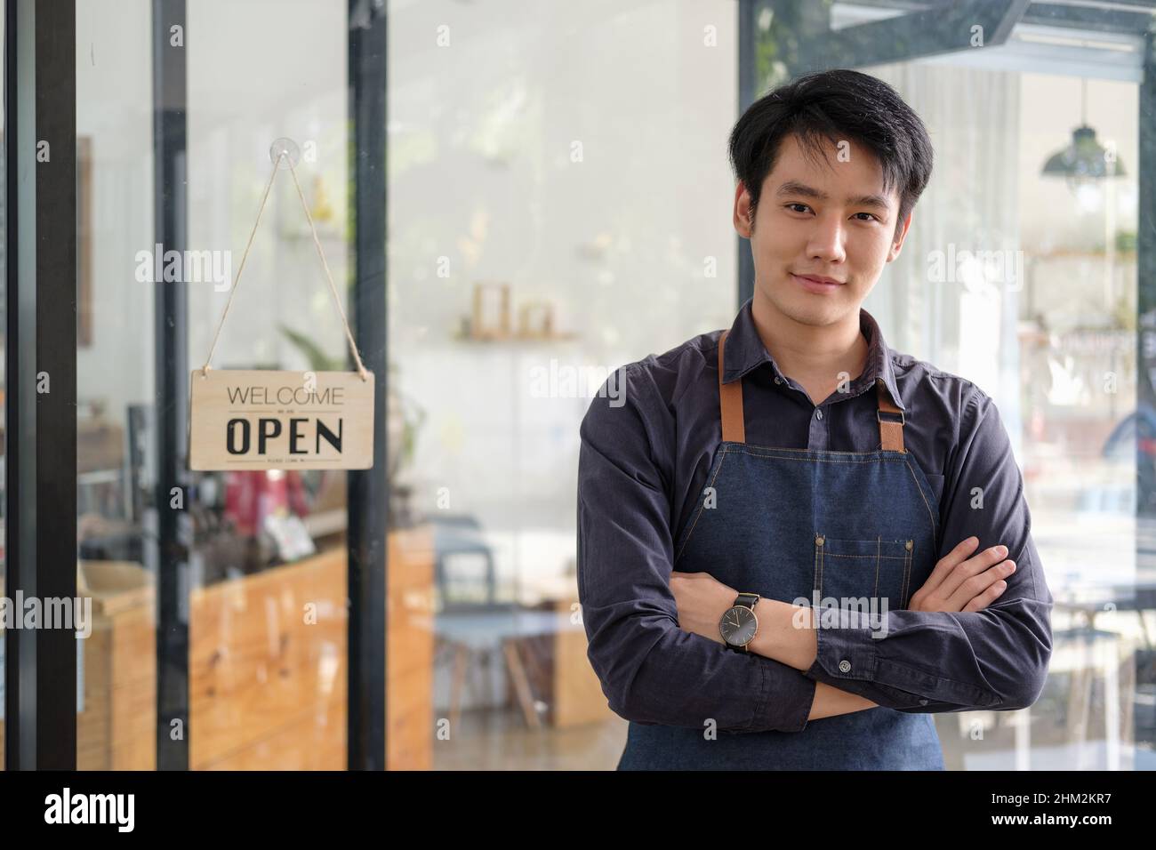 Smarter asiatischer junger Barista, der auf der Schürze ein Tablet hält und vor der Tür des Cafés mit offenem Schild steht. Unternehmer Startup SME Stockfoto