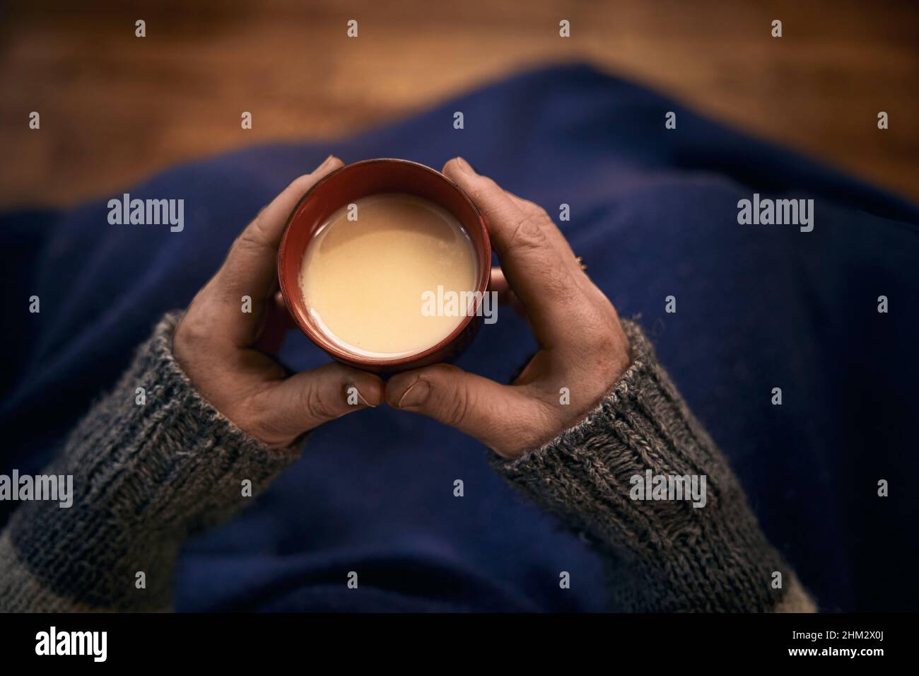 Eine Frau sitzt, bedeckt ihre Beine mit einer blauen Decke und hält eine Tasse heißen Tee mit Milch in den Händen. Stockfoto