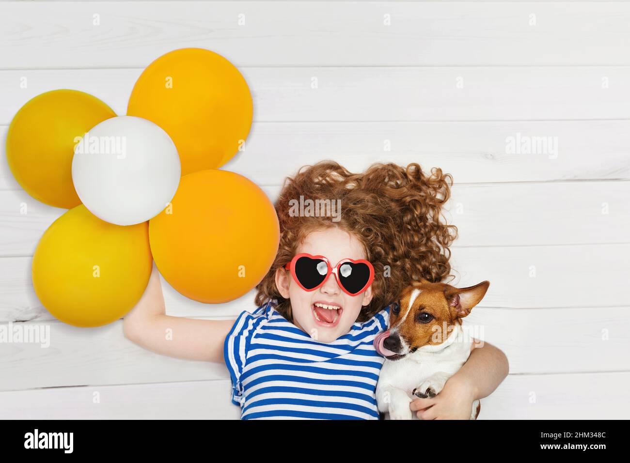 Nettes Mädchen umarmt ihren Freund Jack russell Hund und hält gelbe Ballons. Baby liegt auf dem Holzboden mit Blick von oben. Stockfoto