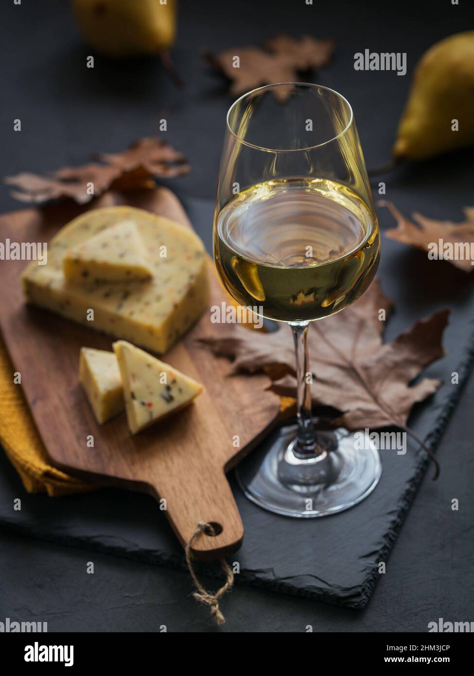 Ein Glas Weißwein wurde mit Käse auf einem Schneidebrett auf dunklem Hintergrund serviert. Herbstpicknick mit Käse, Wein und trockenen Blättern im rustikalen Stil. Stockfoto