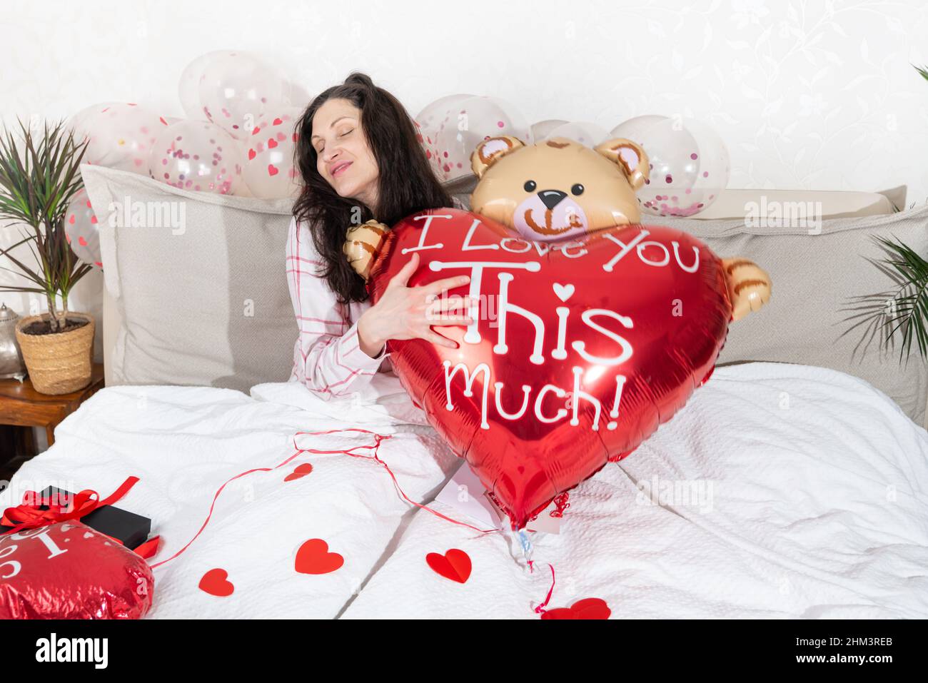 Eine schöne Frau im rosa Schlafanzug mit einem herzförmigen Ballon liegt zu Hause auf dem Bett. Frau am Valentinstag, Geburtstag oder Jahrestag. Liebessymbol Stockfoto