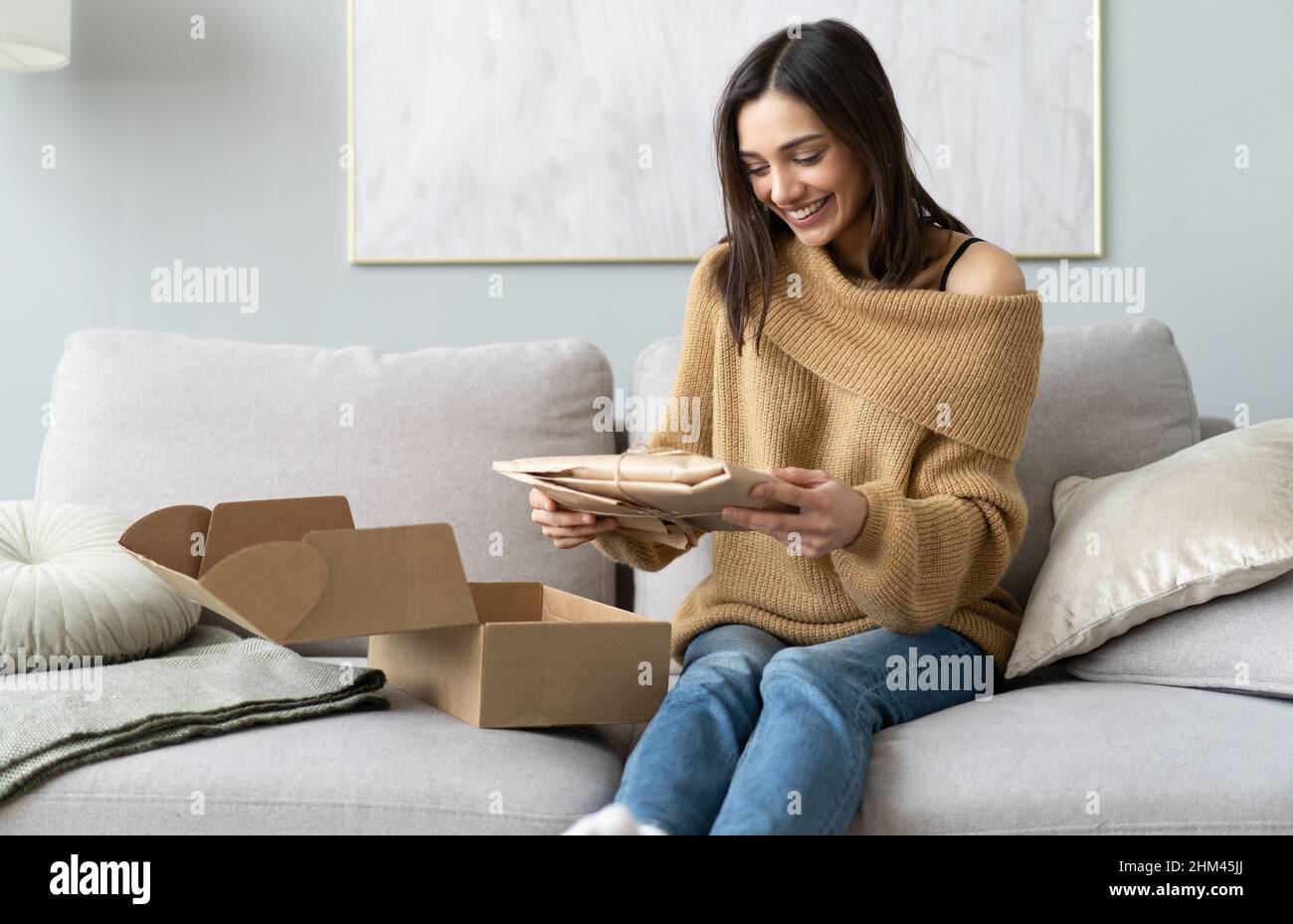 Lächelnd glücklich junge Frau sitzt auf der Couch im Wohnzimmer, Auspacken Karton Paket online bestellen aus dem internationalen Internet-Shop, zufrieden mit Stockfoto