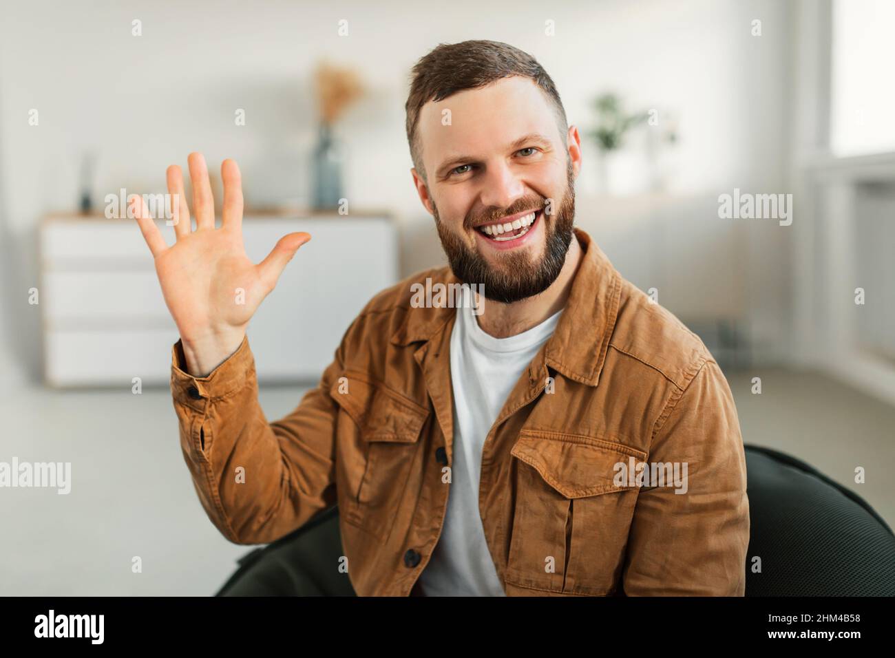Fröhlicher Mann Winkt Mit Der Hand Lächelnd Zur Kamera, Die Zu Hause Sitzt Stockfoto