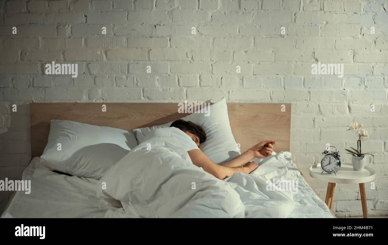 Mann, der unter einer weißen Decke in der Nähe des Vintage-Weckers und einer Orchidee auf dem Nachttisch schläft Stockfoto