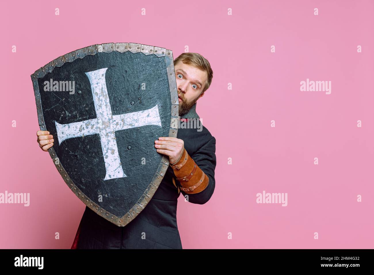 Porträt eines unheimlichen Mannes, eines mittelalterlichen Kriegers oder Bogenschützen in Rüstung, der sich hinter einem Schild versteckt, der über einem rosa Studiohintergrund isoliert ist Stockfoto