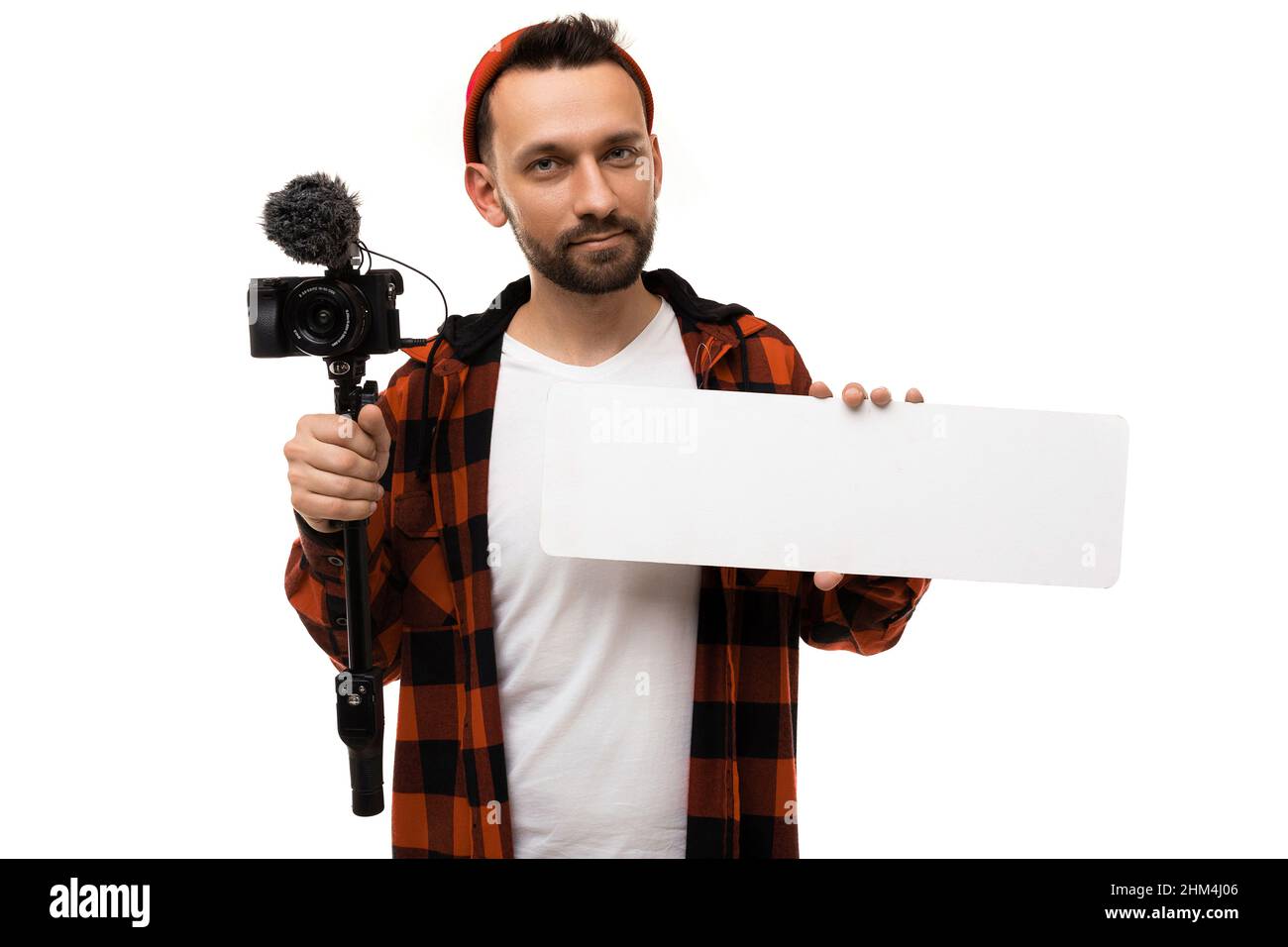 Ein Mann blogger in einem weißen T-Shirt und Hemd in einem rot-schwarzen Käfig mit einer Videokamera und einem Mikrofon blickt in die Linse und hält ein Schild mit Stockfoto