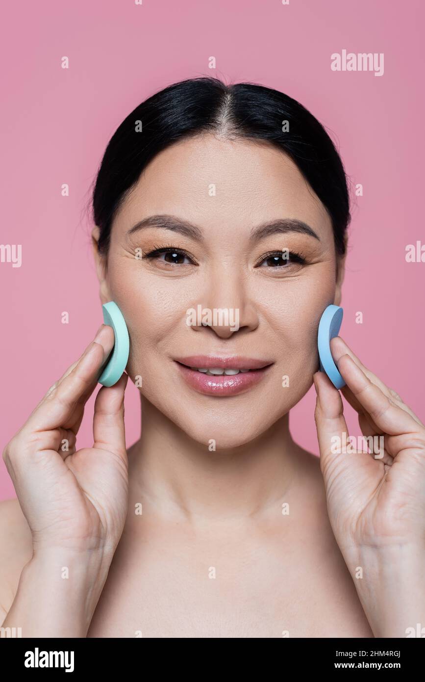 Lächelnd asiatische Frau Reinigung Gesicht mit Schwämmen isoliert auf rosa Stockfoto