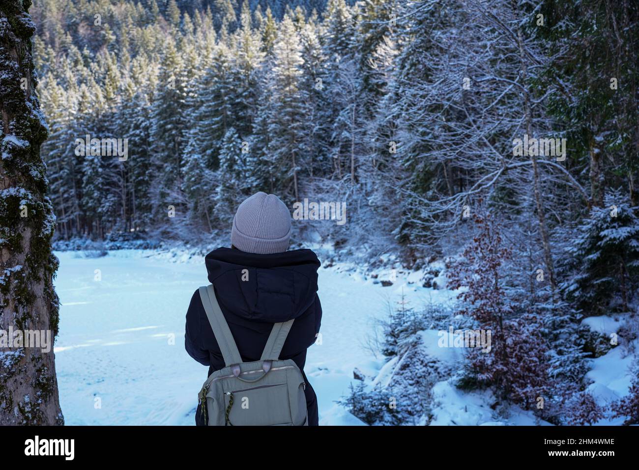 Eine Frau mit Rucksack steht am Ufer des gefrorenen Eibsee und schaut auf schneebedeckte Tannenbäume. Stockfoto