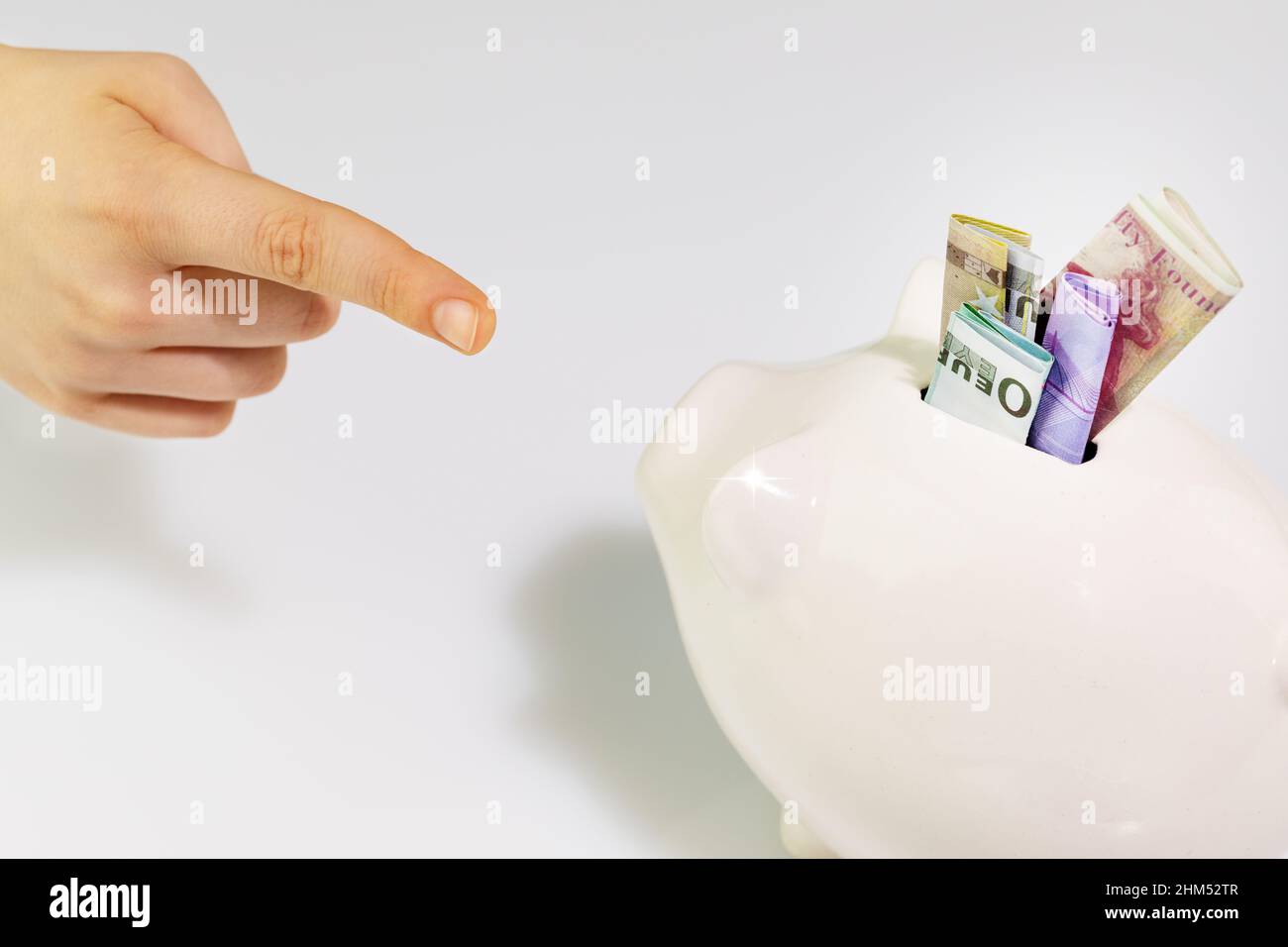 Eine Hand mit ausgestrecktem Finger zeigt auf ein weißes Sparschwein mit Banknoten. Einsparungs- und Investitionskonzept. Weichfokus Stockfoto