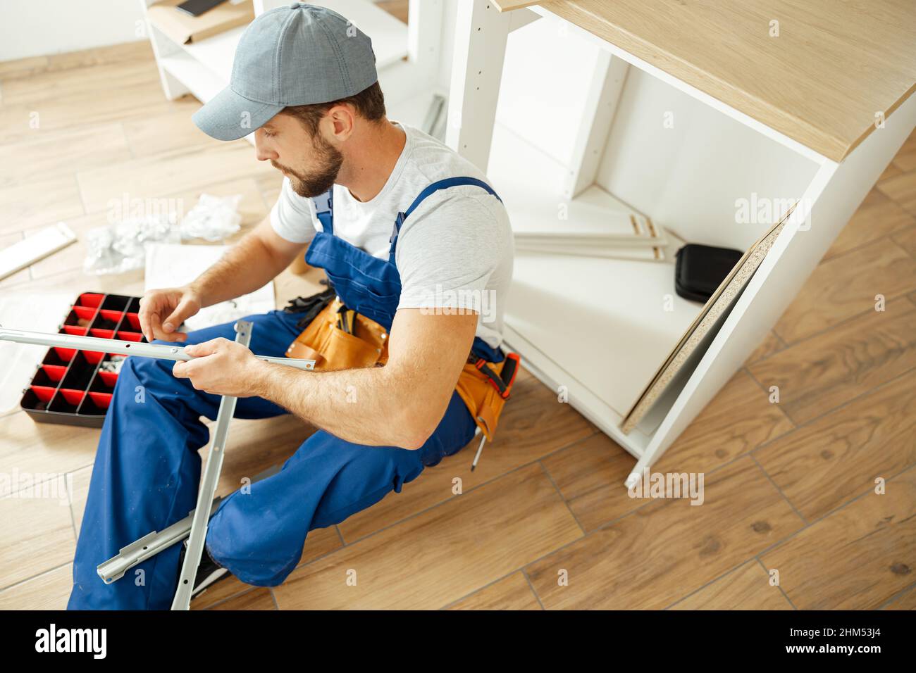 Blick aus der Perspektive auf einen konzentrierten Handwerker in Uniform, der auf dem Boden sitzt, während er in der Küche eine Schublade installiert Stockfoto
