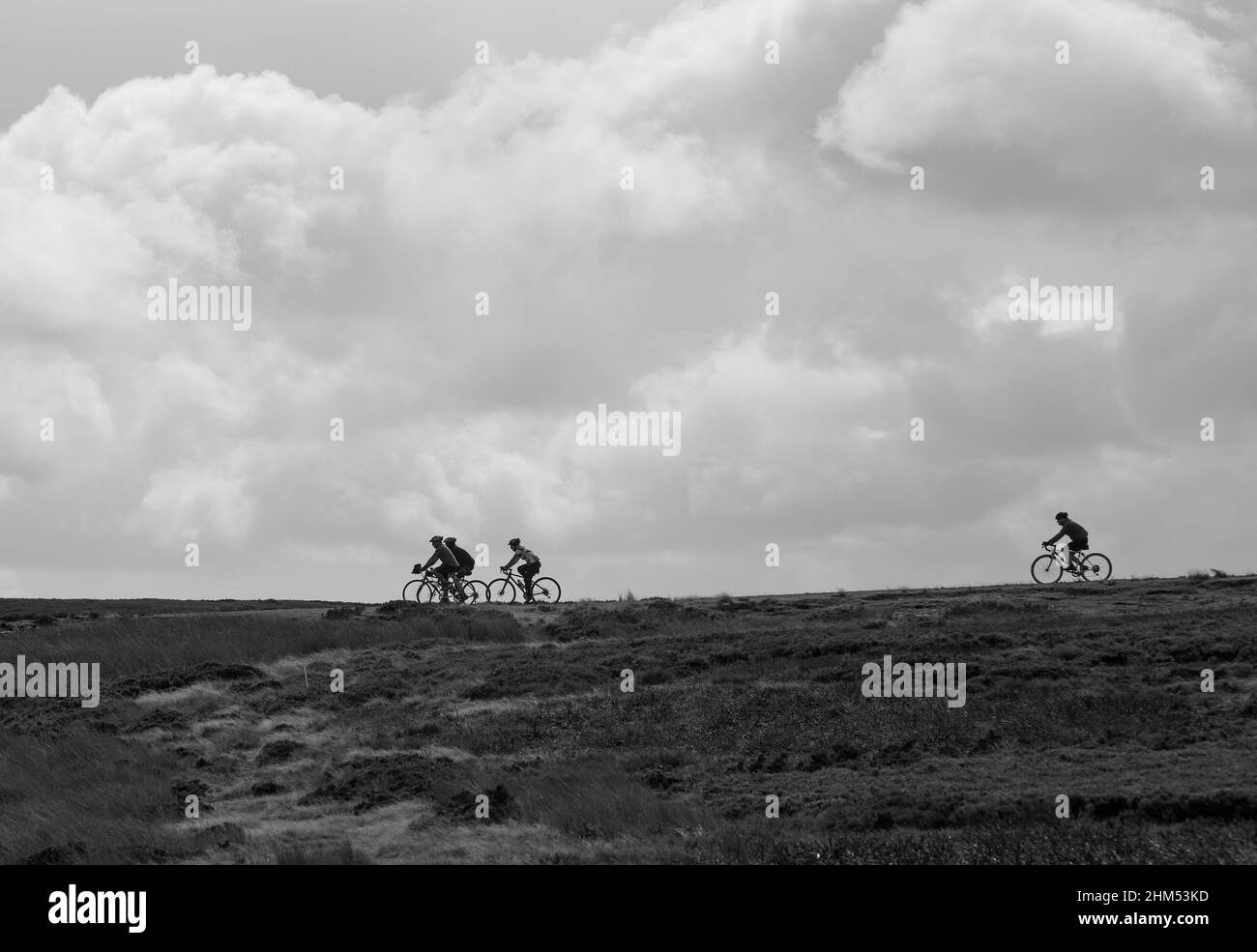 Schwarz-weiß-Bild von vier Radfahrern in Silhouette und am Horizont, während sie durch offene Moorlandschaft mit einem Hintergrund aus Wolken radeln Stockfoto